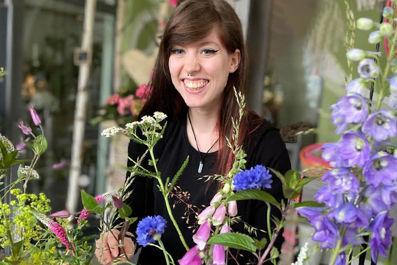  Portrait einer jungen Frau mit dunklen, langen Haaren und schwarzer Kleidung, im Vordergrund verschiedene Blüten