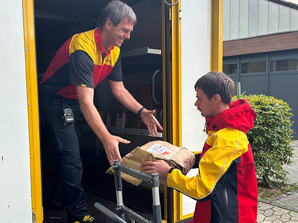 Zwei Männer reichen sich ein Paket. Sie arbeiten beide beim Paketdienst DHL, was an ihrer Kleidung zu erkennen ist.