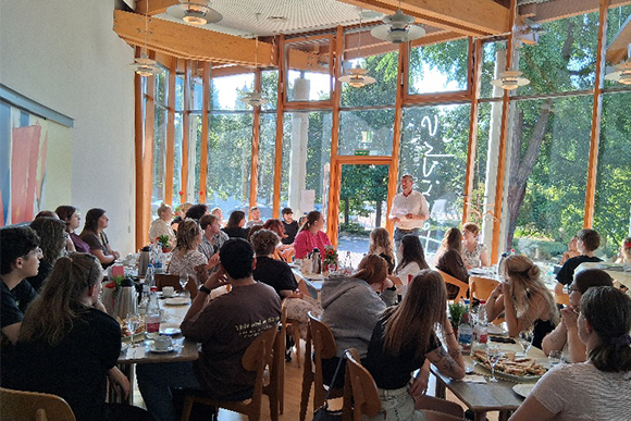 viele junge Menschen sitzen an verschiedenen Tischen im Café, vor ihnen steht ein Mann und hält eine Rede