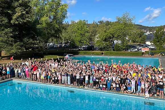 Gruppenfoto mit sehr vielen Menschen, die zwischen zwei Schwimmbecken stehen und die Hände in die Höhe recken