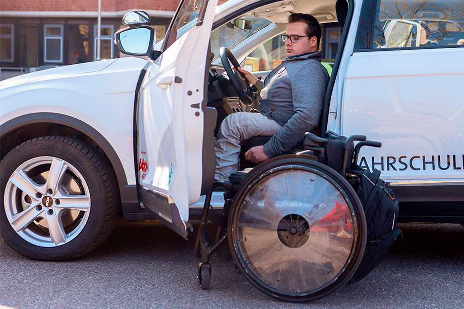 Ein junger Mann sitzt in einem Fahrschulauto. Vor dem Auto steht sein Rollstuhl.
