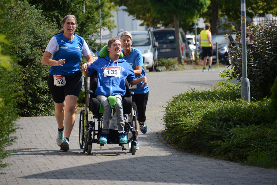 Zwei Läuferinnen im blauen Trikot begleiten einen jungen Mann in blauer Jacke im Rollstuhl bei dem sogenannten "Jedermannslauf".

