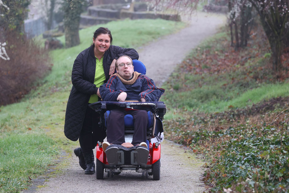 Eine junge Frau geht mit einem jungen Mann im E-Rollstuhl in einem Park spazieren. Beide lächeln in die Kamera.
