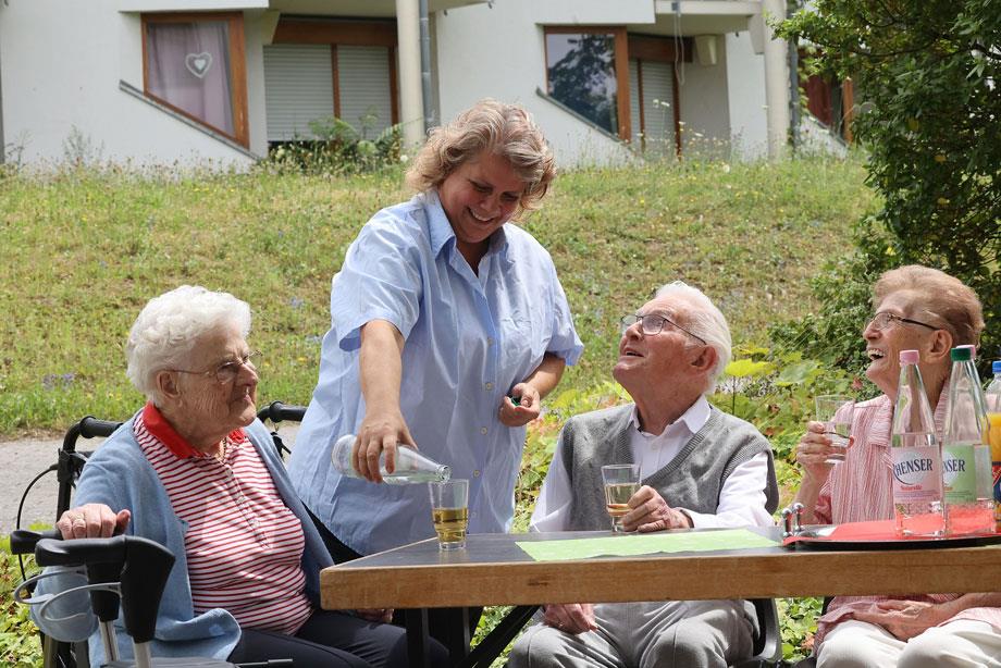 Eine Pflegerin des Seniorenzentrums steht in der Mitte von drei SeniorInnen und schenkt Apfelsaft in ein Glas ein.