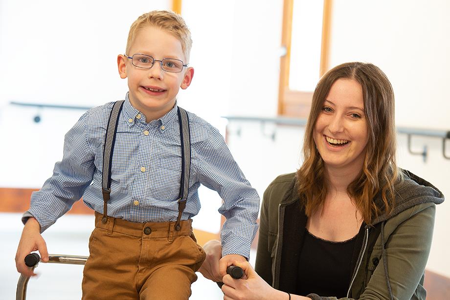 Eine junge Frau steht bei einem kleinen Jungen mit Brille und Rollator. Beide lächeln in die Kamera.