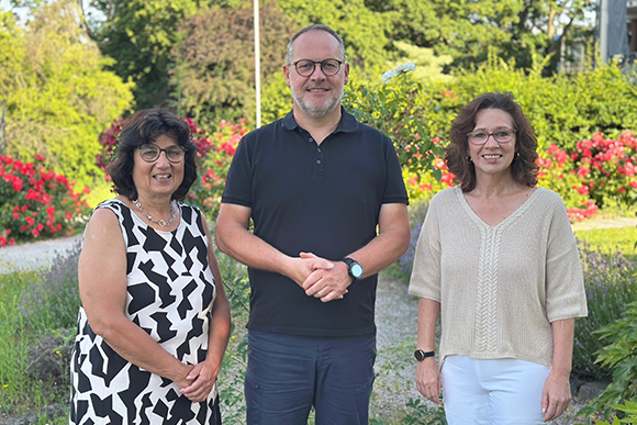 zwei Frauen und ein Mann in der Mitte stehen in einem grünen Garten mit roten Blüten im Hintergrund