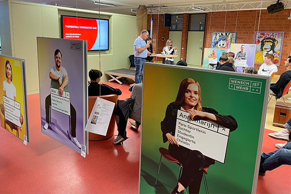 Im Vordergrund hängen drei große Fotos von Menschen vor farbigen Hintergründen, mit Schildern, auf denen steht, wer sie alles sind. Im Hintergrund findet ein Gruppengespräch statt